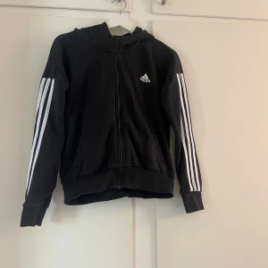 Svart hoodiejacka från Adidas - Svart hoodiejacka från Adidas med dragkedja och klassiska vita ränder längs ärmarna. Jackan har huva, två fickor framtill och Adidas-logga på bröstet. Perfekt för en sportig stil.