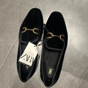 svarta loafers från Zara - Säljer ett par svarta loafers från Zara i mjuk sammet med guldfärgade metalldetaljer framtill. Skorna har en klassisk design med rund tå och låg profil, perfekta för en stilren look.