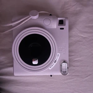 Rosa instantkamera från Instax Mini - Säljer en vit Instax Mini instantkamera med rund lins och inbyggd blixt. Kameran har en enkel design, handledsrem och tydliga reglage för att ta direktbilder. Perfekt för att fånga ögonblick direkt på papper.