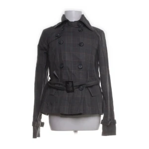 Grå rutig trenchcoat från Cubus - Säljer en grå rutig trenchcoat från Cubus i storlek 34. Jackan har dubbelknäppning, bälte i midjan och långa ärmar med spännen vid handlederna. Perfekt för dig som gillar klassisk stil med modern twist.
