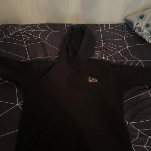 Svart hoodie från Emporio Armani EA7 - Säljer en svart hoodie från Emporio Armani EA7 med logga på bröstet och ärmen. Klassisk huva med dragsko och känguruficka framtill. Perfekt för en stilren och avslappnad look. Passar jätte bra till S och M 