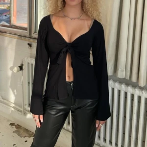 Svart långärmad topp med knyt framtill - Säljer Maddy blouse från Design by Si i svart. Trendig svart långärmad topp med djup v-ringning och knyt framtill som ger en croppad look. Perfekt för att skapa en snygg och edgy outfit. Tillverkad i ett mjukt och stretchigt material. 