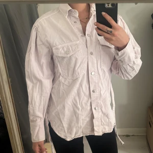 Ljuslila skjorta med bälte - Säljer en ljuslila skjorta med knappar framtill och två bröstfickor. Skjortan har lång ärm och medföljande bälte i midjan för en snygg passform. Perfekt att styla både öppen och stängd.