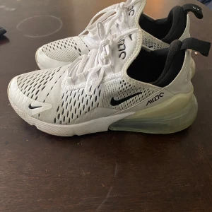 Nike Air Max 270 vita sneakers - Nike Air Max 270 sneakers i vitt med svarta detaljer och stor synlig luftbubbla i hälen. Ovandel i mesh med snörning och svart Nike-logga på sidan. Skorna har en sportig och modern design, perfekt för dig som gillar streetwear.