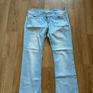 Ljusblå bootcut jeans med dubbla knappar - Säljer ett par ljusblå jeans med bootcut passform och dubbla knappar i midjan. Jeansen har klassiska fickor fram och bak, samt dekorativa sömmar och detaljer. Perfekta för en avslappnad och trendig look.