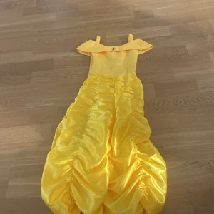 Gul Bell klänning från JerrisApparel - Säljer en gul långklänning från JerrisApparel med axelbandslös design och vackra rynkade detaljer. Klänningen har en glansig satinliknande yta och är perfekt för maskerad. Klassisk prinsesskänsla med elegant form.