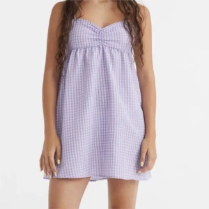 Lila rutig kortklänning - Supersöt lila och vit rutig kortklänning med söt v-ringning och lätt rynk vid bysten. Klänningen är ärmlös och har en avslappnad passform, perfekt för varma dagar.