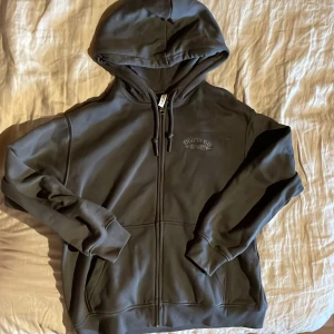 Mörkgrå hoodie från H&M Divided - Säljer en mörkgrå hoodie från H&M Divided med dragkedja och huva. På bröstet finns broderad text 'ATHLETIC CLUB SPORT EST 1996'. Perfekt för en avslappnad stil.