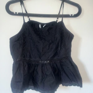 Svart linnetopp med spetsdetaljer - Säljer en svart linnetopp med tunna axelband och spetsdetaljer. Toppen har volang nertill och broderade mönster framtill. Perfekt för sommardagar eller festival.