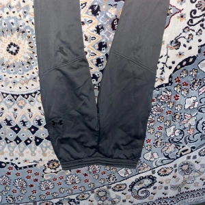 Mörkgrå träningstights från Under Armour - Säljer ett par mörkgrå träningstights från Under Armour med logga på benet. De har bred resår i midjan med tryckt mönster och är tillverkade i ett stretchigt syntetmaterial som passar perfekt för träning.