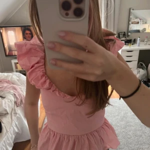 Rosa volangtopp med puffärmar - Supersöt rosa topp med volangdetaljer och puffiga axlar. Djup rundad ringning och volang nertill ger en romantisk känsla. Perfekt att styla med jeans eller kjol för en trendig look.