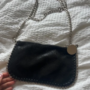 Svart axelväska med kedja - Säljer en svart axelväska med silverfärgad kedja och dekorativ kant i metall. Väskan har dragkedja upptill och en rund metallbricka som detalj. 