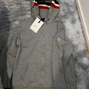 Moncler maglia hoodie - Säljer en grå hoodie från Moncler med dragkedja framtill och huva med ränder i mörkblå, vitt och rött. Tröjan har fickor på sidorna och Moncler-logga på bröstet. Perfekt för en avslappnad och stilren look passar M.