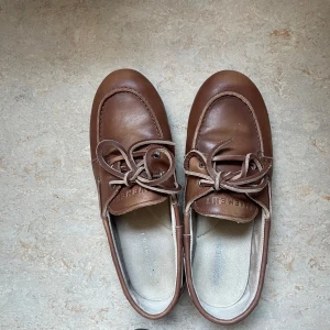 Bruna loafers från Pavement - Säljer ett par klassiska bruna loafers från Pavement i skinn med snörningsdetalj framtill. Skorna har rund tå och platt sula, perfekta för en stilren look. Passar dig som gillar tidlös design och vill ha något som funkar till många outfits. Använda 1 dag. Strl 38. Har kartong kvar. Nypris 1600kr