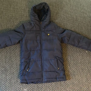 Mörkblå pufferjacka från Lyle & Scott - Säljer en mörkblå pufferjacka från Lyle & Scott Junior med huva och dragkedja. Jackan har två fickor med dragkedja och den klassiska gula loggan på bröstet. Perfekt för kyligare dagar. Storlek Xs/S 14/15 åringar.