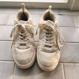 Vita/ beiga sneakers från Champion - Säljer ett par vita sneakers från Champion med klassisk design och snörning. Skorna har rund tå, platt sula och är tillverkade i syntetmaterial med diskret logga på sidan. Perfekta för en clean och sportig stil.