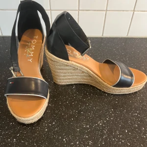 Svarta kilklackssandaler från Tommy Hilfiger - Säljer ett par snygga svarta sandaler från Tommy Hilfiger med hög kilklack i rep och öppen tå. Skorna har bred rem över tårna och justerbart band runt vristen. Perfekta för sommaren och ger en stilren look.