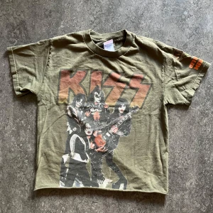 Olivgrön KISS t-shirt - Säljer en olivgrön t-shirt med stort KISS-tryck och bandmedlemmarna på framsidan. Jag har klippt av den där nere för lite med cropped look. På baksidan står det 'Loud and Proud Since 1975' och på ärmen finns texten 'Official Member'. Perfekt för dig som älskar rock och vill sticka ut.