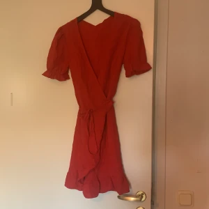 Röd omlottklänning från SHEIN - Säljer en röd omlottklänning från SHEIN med knytband i midjan och volangdetaljer nertill. Klänningen har korta puffärmar och en v-ringad omlottfront. Perfekt för sommarens tillfällen!