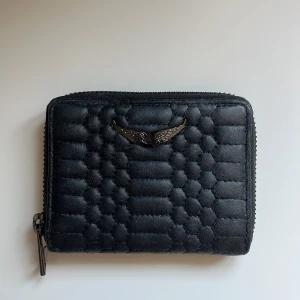 Svart quiltad plånbok från Zadig & Voltaire - Säljer en svart quiltad plånbok från Zadig & Voltaire med coolt vingemblem i metall framtill. Plånboken har dragkedja runtom och flera fack inuti för kort och mynt. Perfekt storlek för att ha med sig överallt.