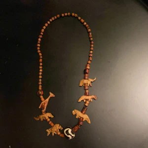 Halsband - Unikt halsband med bruna träpärlor och handgjorda träfigurer av olika djur, bland annat giraff och elefant. Halsbandet har även en större pärla med svartvitt mönster. Perfekt för dig som gillar naturinspirerade accessoarer.