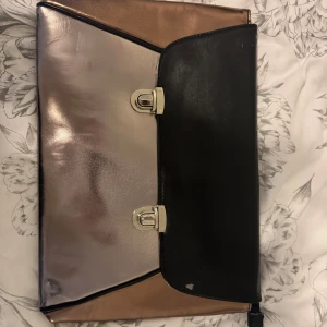 Metallic kuvertväska från Ellos Accessoriesi - Säljer en snygg kuvertväska i metallic från Ellos Accessories. Väskan har en mix av roséguld och svart med silvriga detaljer och dragkedja på baksidan. Perfekt för att bära det viktigaste och stängs med två spännen framtill.