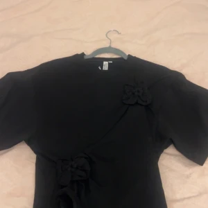 Svart t shirt med rosettdetaljer från & Other Stories - Svart t shirt från & Other Stories med fina rosettdetaljer framtill och vid axeln. Klänningen har korta ärmar och en elegant, enkel design som passar perfekt till olika tillfällen.