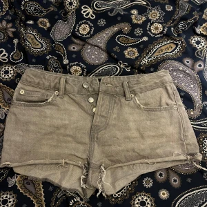 Gråa jeansshorts med fransar - Säljer ett par gråa jeansshorts med råa franskantade benslut och klassisk femficksdesign. Shortsen har låg midja och dubbla knappar framtill. Perfekta för sommardagar! 