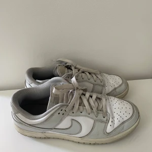 Vita Nike Dunk sneakers - Säljer ett par klassiska vita Nike Dunk sneakers med grå detaljer. Skorna har snörning, perforerad tå och platt sula. Perfekta för dig som gillar en stilren och sportig look. Ikonisk Nike-logga på sidan och sulan.