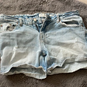 Ljusblå jeansshorts från C Denim - Ljusblå jeansshorts från C Denim med råa kanter och klassisk femficksdesign. Shortsen har normal passform och är tillverkade i denim med slitna detaljer för en avslappnad look. Perfekta för varma dagar.