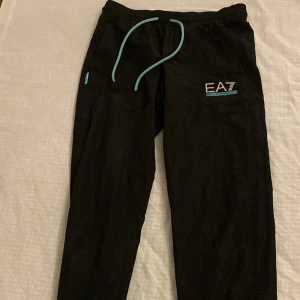Svarta mjukisbyxor från Emporio Armani EA7 - Säljer ett par svarta mjukisbyxor från Emporio Armani EA7 med turkosa snören och logga på benet. Byxorna har normal passform och resår i midjan. Perfekta för chill eller träning. Storleken är S och xs 