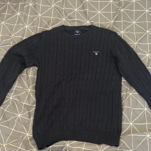 Mörkblå kabelstickad tröja från GANT - Säljer en klassisk mörkblå kabelstickad tröja från GANT i 100% bomull. Tröjan har rund halsringning, ribbade muddar och diskret GANT-logga broderad på bröstet. Perfekt för en stilren och tidlös look.