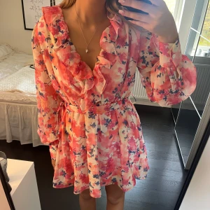 Blommig volangklänning - Superfin kortklänning med blommigt mönster i rosa, rött och vitt. Klänningen har långa ärmar, volangdetaljer längs ringningen. Aldrig använd och prislappen kvar 