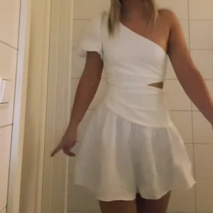 studentklänning - Säljer en vit kortklänning med one shoulder-design och volangdetalj på axeln. Klänningen har en asymmetrisk skärning och liten cut-out i midjan. Endast provad