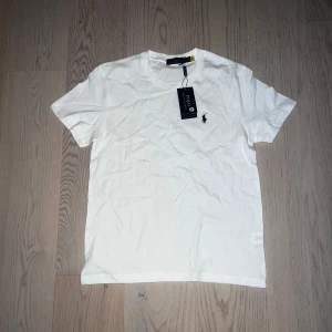 Vit t-shirt från Polo Ralph Lauren - Säljer en klassisk vit t-shirt från Polo Ralph Lauren med den ikoniska broderade loggan på bröstet. T-shirten har rund hals och korta ärmar, perfekt för en stilren och enkel look.