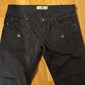 Svarta byxor från Fornarina med fickdetaljer - Säljer ett par svarta lågmidjade byxor från Fornarina med delorstiva fickor framtill och knappar i mässingsfärg. Stuprörsjeans med dragkedja nedtill vid benen.