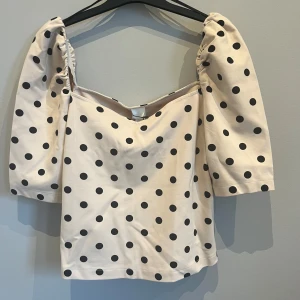 Beige topp med svarta prickar och puffärmar - Trendig beige topp med svarta prickar och stora puffärmar. Toppen har en fyrkantig ringning.