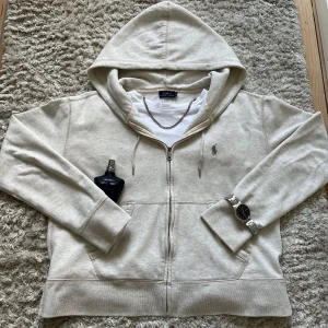 Ralph Lauren Zip Up - Säljer en stilren beige hoodie från Polo Ralph Lauren med dragkedja och klassisk logga broderad på bröstet. Den är storlek M (barnstorlek) men passar XS i herrstorlek. Tröjan har huva med dragsko och två fickor framtill. Perfekt för alla outfits med en stilren look.