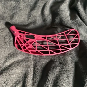 Rosa innebandyblad - Säljer ett rosa innebandyblad med öppen, gallerliknande design. Bladet är böjt och har en lätt konstruktion i plast, perfekt för snabba skott och bra bollkontroll. Passar de flesta standardklubbor för innebandy.