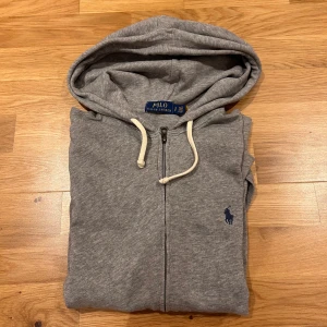 Grå hoodie från Polo Ralph Lauren - Säljer en klassisk grå hoodie från Polo Ralph Lauren med dragkedja och vit dragsko i huvan. Tröjan har en liten blå logga broderad på bröstet och är tillverkad i mjuk bomull. Perfekt för en avslappnad stil. Lite använd. Ny pris 2 000 kr, pris kan diskuteras 