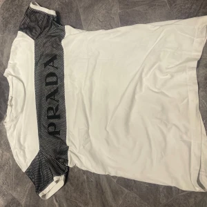 Vit t-shirt från Prada med svarta detaljer - Säljer en vit t-shirt från Prada med svarta partier och stort PRADA-tryck över axlarna. T-shirten har rund halsringning och korta ärmar. Perfekt för dig som gillar stilrena märkesplagg.