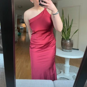 Vinröd långklänning med oneshoulder och öppen rygg - Säljer en vinröd långklänning med oneshoulder och snygg öppen rygg med knyt. Klänningen har en elegant och figurnära passform, perfekt för bal eller fest. Materialet är glansigt och följsamt, vilket ger en lyxig känsla.