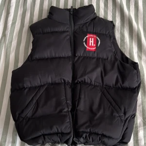 Svart dunväst från Hoodrich - Svart dunväst från Hoodrich med hög krage och dragkedja framtill. Västen har tryckt logga och text både fram och bak samt två stora fickor. Perfekt för lager-på-lager-stil under kyliga dagar.