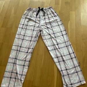 Rosa och vita rutiga mjukisbyxor med snörning - Säljer ett par mjuka och sköna mjukisbyxor i ljusrosa med mörkblått rutmönster. Byxorna har resår i midjan och svart snörning för justerbar passform. Perfekta för en avslappnad stil hemma.