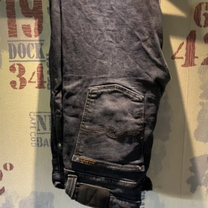 Svarta jeansbyxor från Nudie Jeans - Säljer ett par svarta jeansbyxor från Nudie Jeans med klassisk femficksdesign och snygga detaljer på bakfickan. Byxorna har normal passform och är perfekta för dig som gillar stilrena och tidlösa jeans.