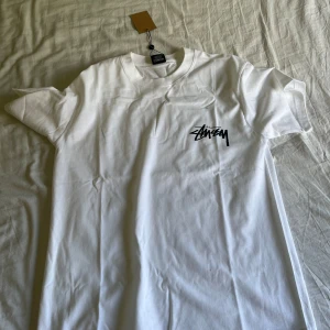 Vit Stüssy t-shirt med tryck - Säljer en vit t-shirt från Stüssy med svart logga på bröstet och stor logga samt rosa tärningar på ryggen. Klassisk passform och korta ärmar. Perfekt för dig som gillar streetwear och vill sticka ut med en snygg detalj på ryggen.