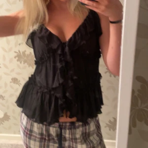 Svart volangig linnetopp med knyt - Supersöt svart linnetopp med volanger och knyt framtill. Tunt, plisserat material och smala axelband. Perfekt för att skapa en trendig och feminin look. Storlek L men sitter som en M/S