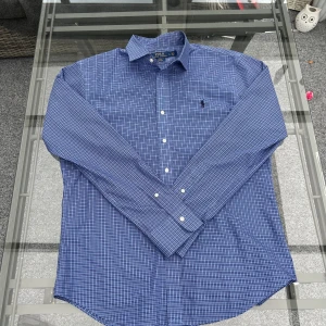 Ralph Lauren blå rutig skjorta – slim fit, strl L - (PRISET DISKUTERBART)                                                                                   Blå rutig skjorta från Ralph Lauren i storlek L – klassisk men ändå modern med slim fit-passform och den ikoniska broderade loggan på bröstet. Perfekt till både vardag och mer dressade tillfällen. Nästan ny och i jättefint skick! Passar dig som gillar stilren, tidlös design med kvalitet.