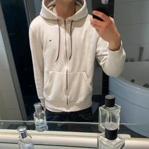 Vit hoodie från Tommy Hilfiger - Säljer en klassisk vit hoodie från Tommy Hilfiger med dragkedja och huva. Tröjan har två fickor framtill, justerbara snören i huvan med Hilfigers signaturfärger och en liten broderad logga på bröstet. Perfekt för en stilren och avslappnad look.