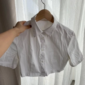 Vit croppad skjorta med kort ärm - Säljer en stilren vit croppad skjorta med korta ärmar och klassisk krage. Skjortan har knappar framtill. Perfekt för en trendig och avslappnad look.
